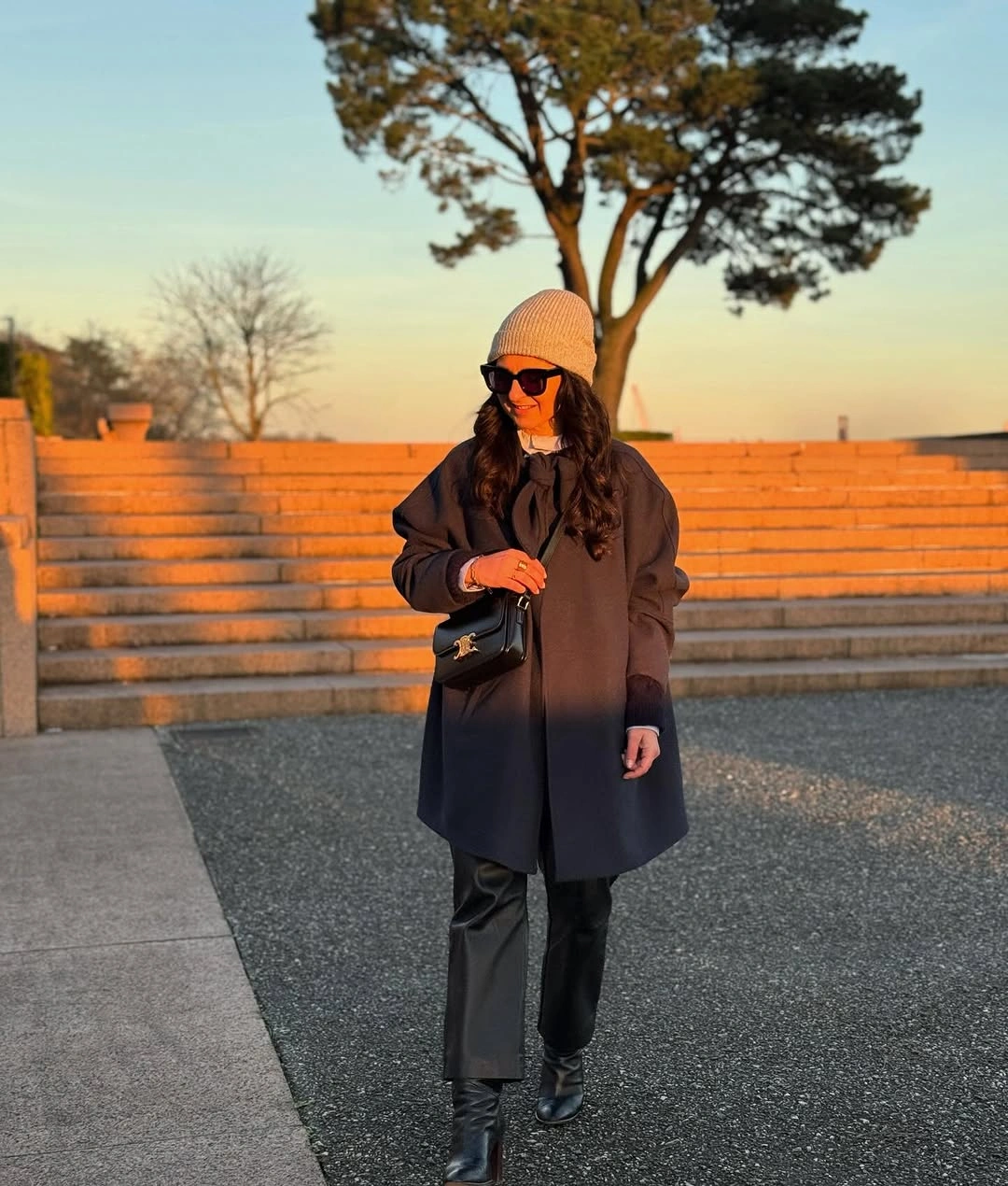 Woman in long dark coat, light beanie, glossy trousers, and pointed boots