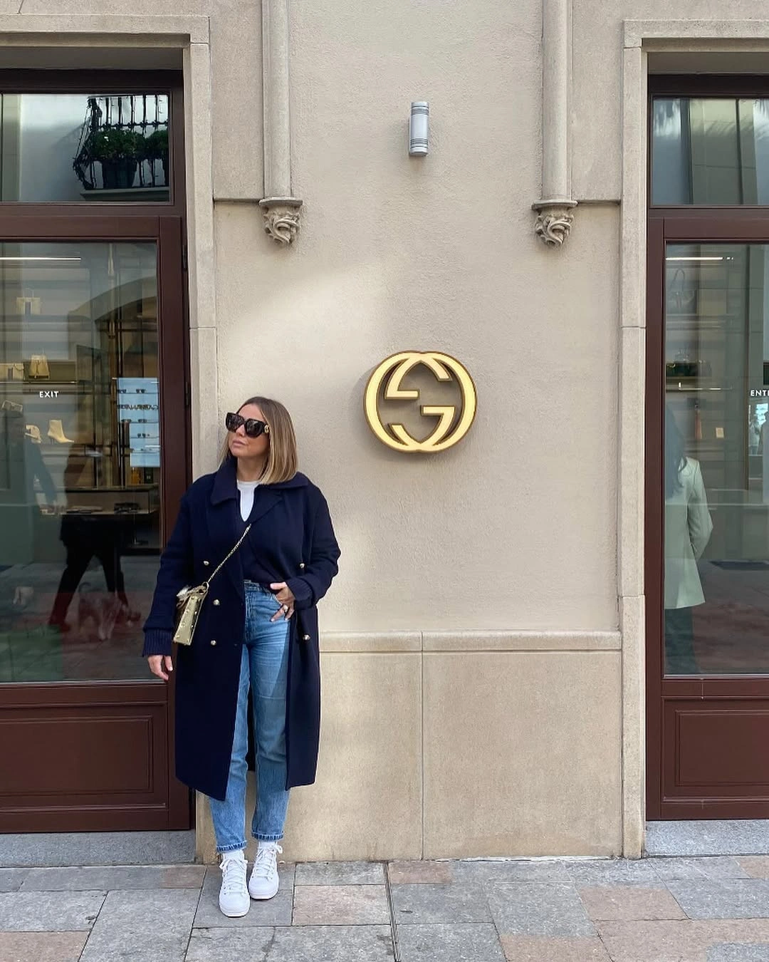 Woman in long navy coat, blue jeans, white sneakers, and sunglasses