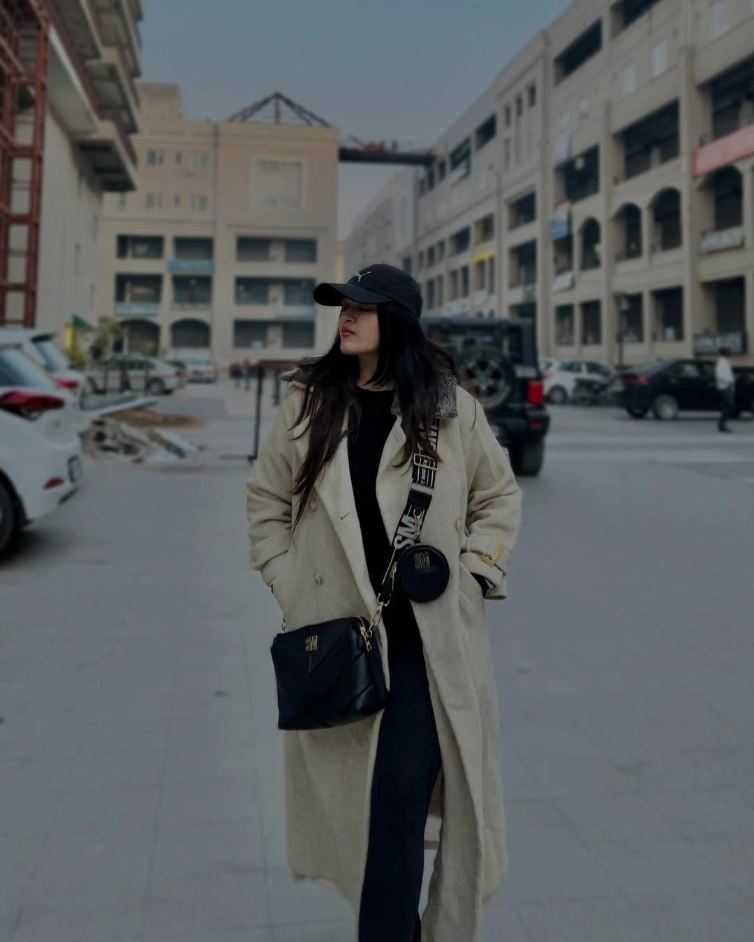 Woman wearing long beige coat, black cap, and crossbody bags outdoors