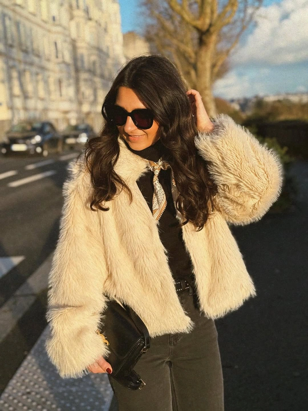 Woman in cropped ivory plush jacket, dark trousers, scarf, and sunglasses