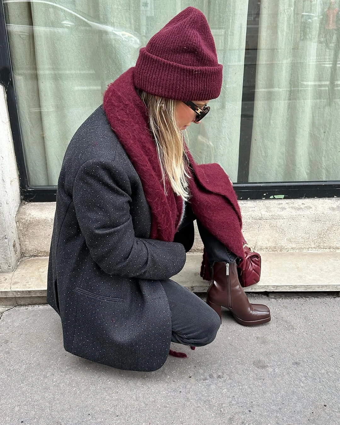 Woman crouching in charcoal jacket and trousers, burgundy scarf, beanie, and ankle boots