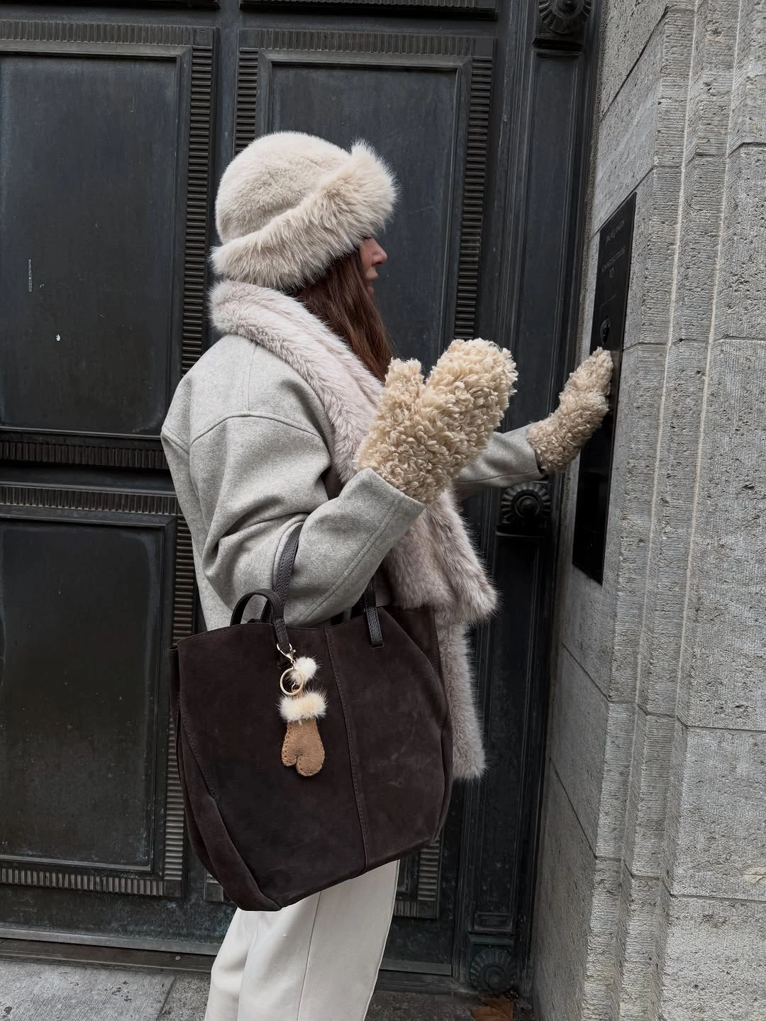 Woman in gray coat, fluffy hat, cream mittens, and dark tote