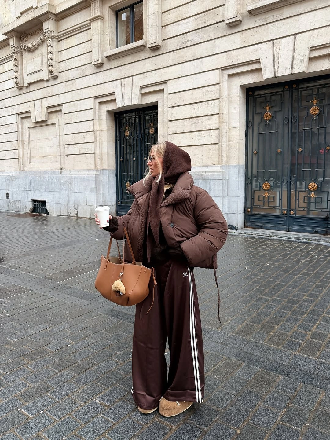 Woman in brown puffer, hooded layer, track pants, and tan tote